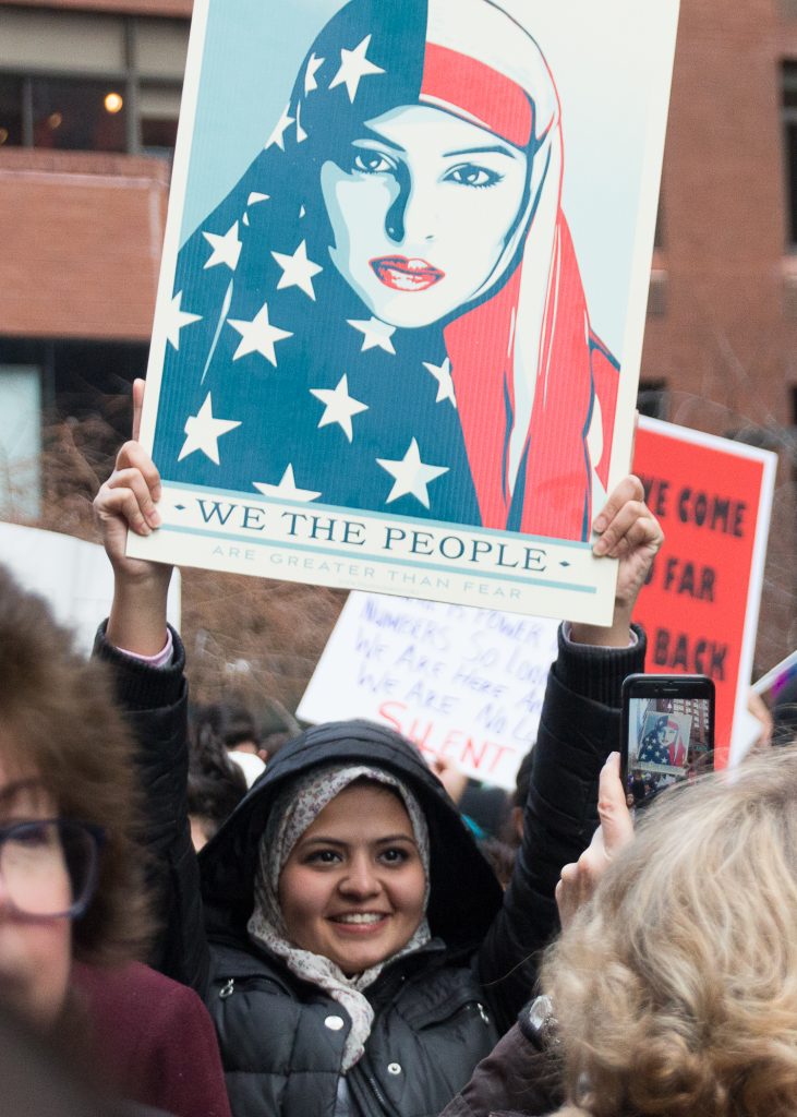 A young woman in a headscarf at the New York City Women's March holds up Shepard Fairey's "WE THE PEOPLE" poster featuring a woman in an American flag headscarf.